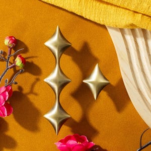 Puede incluir: Tres tiradores de armario en forma de estrella de color latón y un solo pomo en forma de estrella se muestran sobre un fondo de color mostaza. Los tiradores tienen un diseño apilado único. Flores decorativas y tela complementan la presentación.