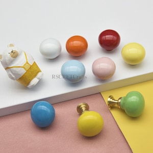 Könnte beinhalten: Eine Sammlung farbenfroher Keramik-Schrankknöpfe und ein bärenförmiges Dekorationselement. Die Knöpfe sind in verschiedenen Farben erhältlich, darunter Weiß, Orange, Rot, Gelb, Blau und Rosa. Der Bär ist weiß mit gelben Latzhosen. RSCRFTCrafts ist aufgedruckt.