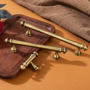 May include: Antique brass cabinet pulls with a classic design. The pulls have a cylindrical bar with decorative end caps and are mounted on small, triangular feet. The hardware is displayed on a wooden tray and a leather surface.