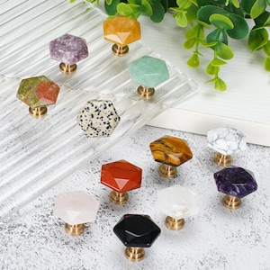Könnte beinhalten: Eine Sammlung von zwölf sechseckigen Schrankknöpfen aus verschiedenen polierten Natursteinen, darunter Amethyst, Tigerauge, Dalmatiner-Jaspis und Quarz, jeweils mit einem goldfarbenen Metallsockel und einer facettierten Oberfläche.