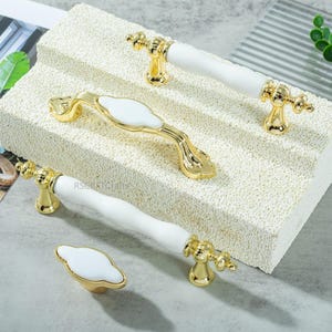 May include: A collection of gold-toned cabinet hardware with white ceramic accents. The set includes various handle styles and a knob, all featuring ornate detailing. The hardware is displayed on a textured, off-white surface.