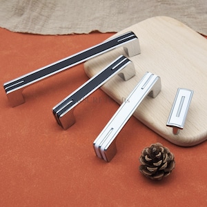 Op de afbeelding: Een verzameling kastbeslag, waaronder drie rechthoekige handgrepen en een kleine knop. De handgrepen hebben een zilveren afwerking met zwarte of witte middenaccenten. Een dennenappel ligt naast de hardware op een houten snijplank.