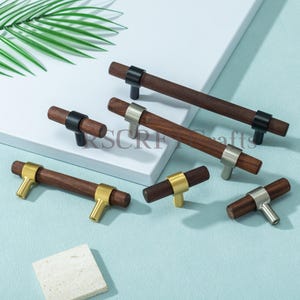 Peut inclure: Plusieurs poignées de meubles en bois avec des accents métalliques variés. Les poignées présentent des poignées en bois brun foncé associées à des ferrures métalliques noires, dorées et argentées. Les ferrures sont en forme de T. Les poignées sont disposées sur une surface bleu clair.