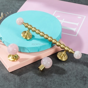 Puede incluir: Herrajes de gabinete de latón con detalles de cuarzo rosa. El conjunto incluye un tirador largo con una serie de esferas de latón y extremos de cuarzo rosa, y dos pomos más pequeños. Los herrajes se muestran sobre un fondo azul y rosa.