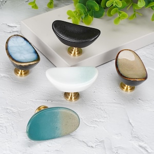 Puede incluir: Cinco pomos de armario de forma ovalada con superficies lisas y redondeadas y bases de metal dorado. Los pomos presentan acabados variados que incluyen negro sólido, blanco brillante, azul claro, beige y un degradado de verde azulado y beige.