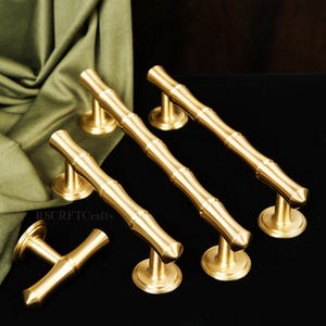 May include: A set of five brass bamboo-style cabinet pulls. The pulls are gold-colored and have a textured, bamboo-like design. They are arranged on a black surface with a draped olive green fabric in the background. The pulls are approximately 6 inches long.