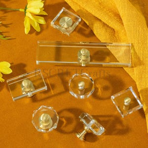 Op de afbeelding: Een verzameling heldere acryl kastknoppen met messing accenten. De knoppen hebben verschillende vormen, waaronder vierkant, rechthoekig en rond. De knoppen worden gepresenteerd op een mosterdkleurig oppervlak met gele bloemen en gele stof.