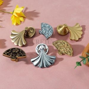 Puede incluir: Una colección de tiradores decorativos para armarios en varios acabados metálicos, incluyendo dorado, plateado y bronce. Los tiradores presentan diseños ornamentados en forma de abanico y florales, añadiendo un toque de elegancia. Los tiradores están dispuestos sobre una superficie rosa.