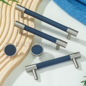 Könnte beinhalten: Ein Set von Schrankbeschlägen, darunter drei Stangengriffe und zwei runde Knöpfe. Die Griffe haben eine gebürstete Metalloberfläche mit marineblauen lederummantelten Griffen. Die Knöpfe haben ebenfalls eine marineblaue Ledermitte.