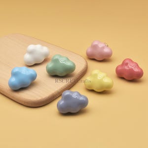 Może przedstawiać: Kolekcja kolorowych, ceramicznych gałek do szafek w kształcie chmur w r&oacute;żnych pastelowych kolorach, w tym białym, niebieskim, zielonym, r&oacute;żowym, ż&oacute;łtym, czerwonym i szarym. Gałki są prezentowane na drewnianej desce do krojenia na ż&oacute;łtym tle.