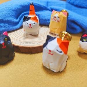 Puede incluir: Una colección de figuras de gatos de madera talladas a mano en varios colores, incluyendo negro, blanco, naranja y amarillo. Cada gato tiene un diseño y una expresión facial únicos. Las figuras se exhiben sobre una superficie de madera.