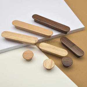 May include: A collection of wooden cabinet hardware, including rectangular pulls and round knobs. The pulls are in light and dark wood tones, and the knobs are a lighter wood. The hardware is displayed on a white and tan surface.