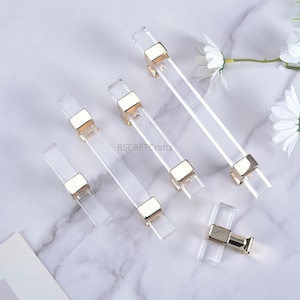 Puede incluir: Tiradores de armario de acrílico transparente con detalles metálicos dorados. Los tiradores son rectangulares con un cuerpo transparente y herrajes de montaje metálicos dorados. La imagen incluye el texto "RSCRFTCrafts".