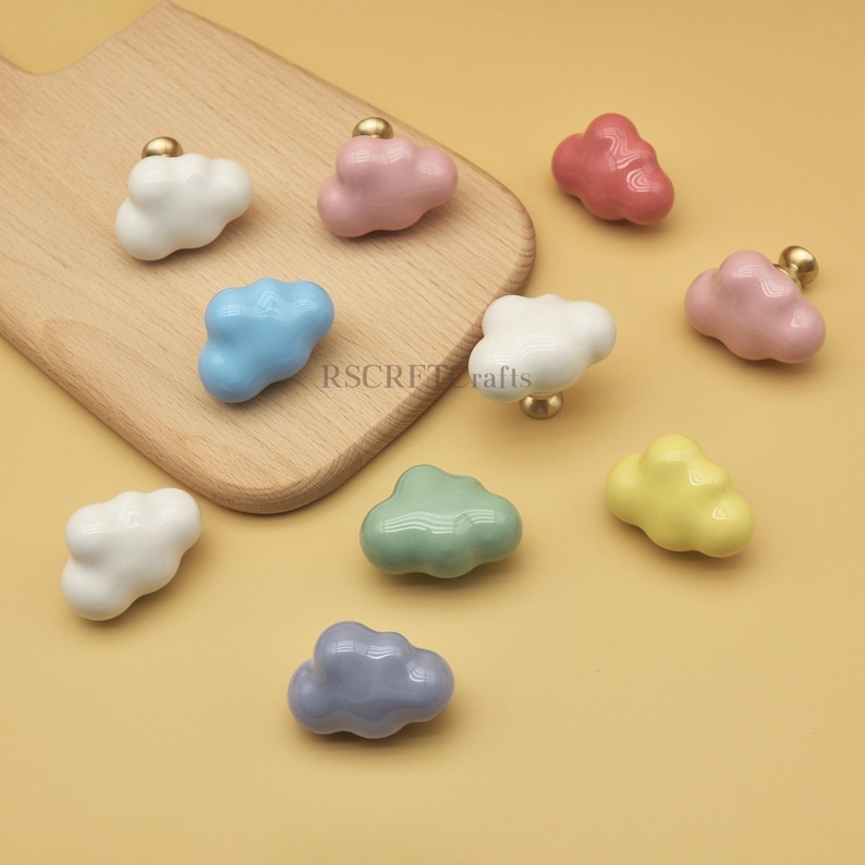 Może przedstawiać: Kolekcja uchwyt&oacute;w do szafek w kształcie chmur w r&oacute;żnych kolorach, w tym białym, r&oacute;żowym, niebieskim, zielonym, ż&oacute;łtym i szarym. Każdy uchwyt ma złotą podstawę. Uchwyty są ułożone na drewnianej desce do krojenia i ż&oacute;łtej powierzchni.