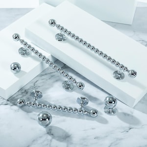 May include: Shiny chrome door handles and knobs. The handles are long, with a series of small, round beads. The knobs are also round. The hardware is displayed on a white surface with a marble background.