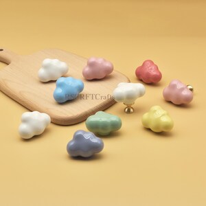 Może przedstawiać: Kolekcja ceramicznych gałek do szafek w kształcie chmur w r&oacute;żnych kolorach, w tym białym, r&oacute;żowym, niebieskim, zielonym, ż&oacute;łtym i szarym. Gałki są prezentowane na drewnianej desce do krojenia i ż&oacute;łtym tle. Widoczny jest tekst "RSRFTCrafts".