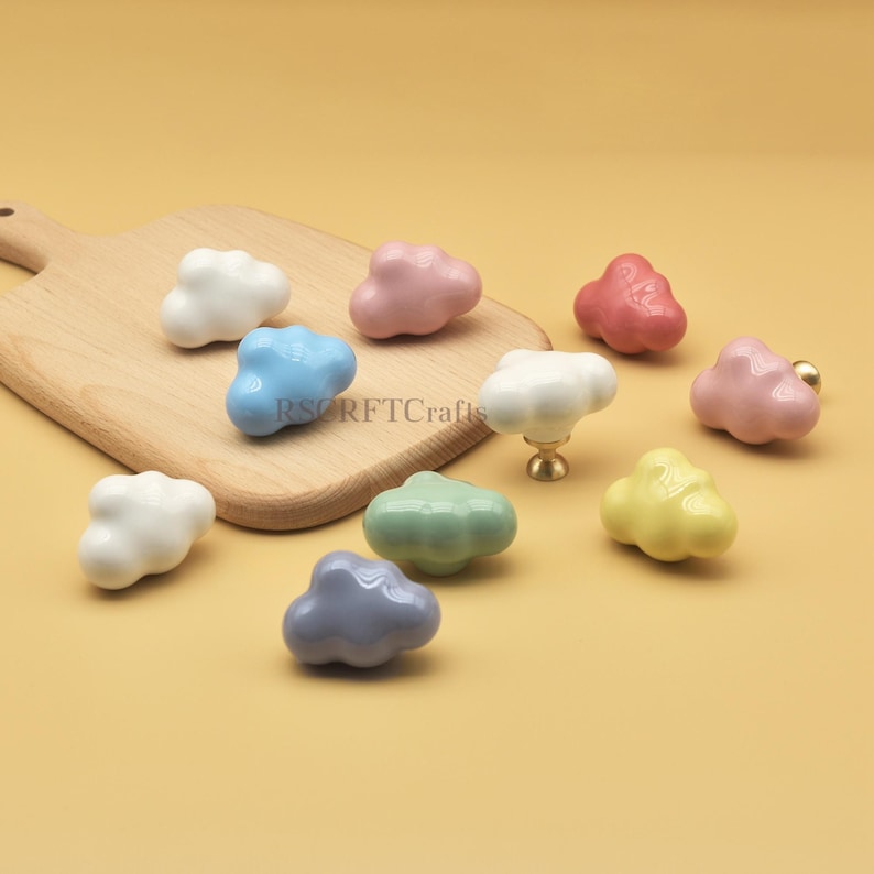 Może przedstawiać: Kolekcja ceramicznych gałek do szafek w kształcie chmur w r&oacute;żnych kolorach, w tym białym, r&oacute;żowym, niebieskim, zielonym, ż&oacute;łtym i szarym. Gałki są prezentowane na drewnianej desce do krojenia i ż&oacute;łtym tle. Widoczny jest tekst "RSRFTCrafts".