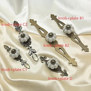 Może przedstawiać: Asortyment gałek i płytek do szafek w stylu antycznym. Każda gałka ma biały, ceramiczny wz&oacute;r w kształcie kwiatu z mosiężnym środkiem. Płytki mają r&oacute;żne ozdobne kształty i wykończenia, z etykietami tekstowymi takimi jak "knob+plate B1" i "knob+plate C2".