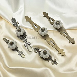 Może przedstawiać: Asortyment uchwyt&oacute;w do szafek w stylu antycznym. Każdy uchwyt posiada białe, kwiatowe pokrętło z mosiężnym środkiem, zamontowane na ozdobnych metalowych płytkach tylnych w r&oacute;żnych wzorach i wykończeniach. Okucia są prezentowane na kremowej tkaninie.