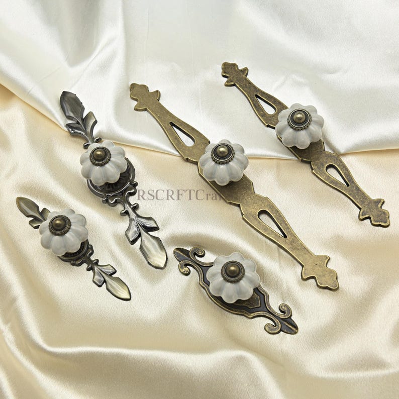 Może przedstawiać: Asortyment uchwyt&oacute;w do szafek w stylu antycznym. Każdy uchwyt posiada białe, kwiatowe pokrętło z mosiężnym środkiem, zamontowane na ozdobnych metalowych płytkach tylnych w r&oacute;żnych wzorach i wykończeniach. Okucia są prezentowane na kremowej tkaninie.