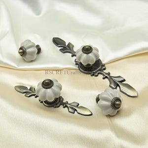 Może przedstawiać: Zestaw białych ceramicznych gałek i uchwyt&oacute;w do szafek z kwiatowym wzorem i antycznym brązowym osprzętem. Gałki i uchwyty są ułożone na kremowej tkaninie. Widoczny jest tekst "RSCRFTcrafts".