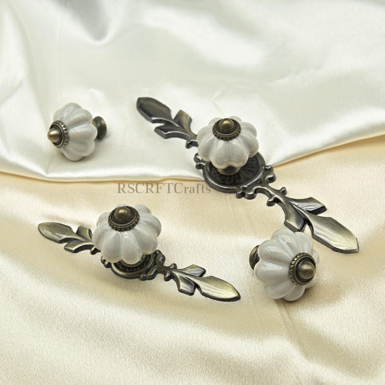 Może przedstawiać: Zestaw białych ceramicznych gałek i uchwyt&oacute;w do szafek z kwiatowym wzorem i antycznym brązowym osprzętem. Gałki i uchwyty są ułożone na kremowej tkaninie. Widoczny jest tekst "RSCRFTcrafts".