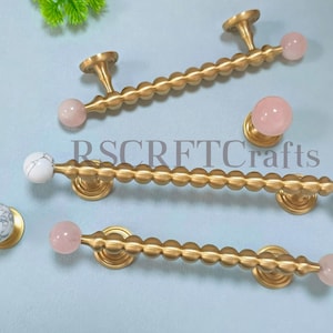 Peut inclure: Quincaillerie de placard en laiton avec des accents de quartz rose et de marbre. L'image présente des poignées et des boutons de tiroir. Les poignées ont un design perlé et mesurent environ 15 cm de long. Les boutons sont ronds.