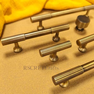 Puede incluir: Un conjunto de herrajes de latón antiguo para armarios, que incluye tiradores y pomos. Los herrajes presentan un diseño acanalado y un acabado cálido y envejecido. Las piezas están dispuestas sobre una superficie neutra.