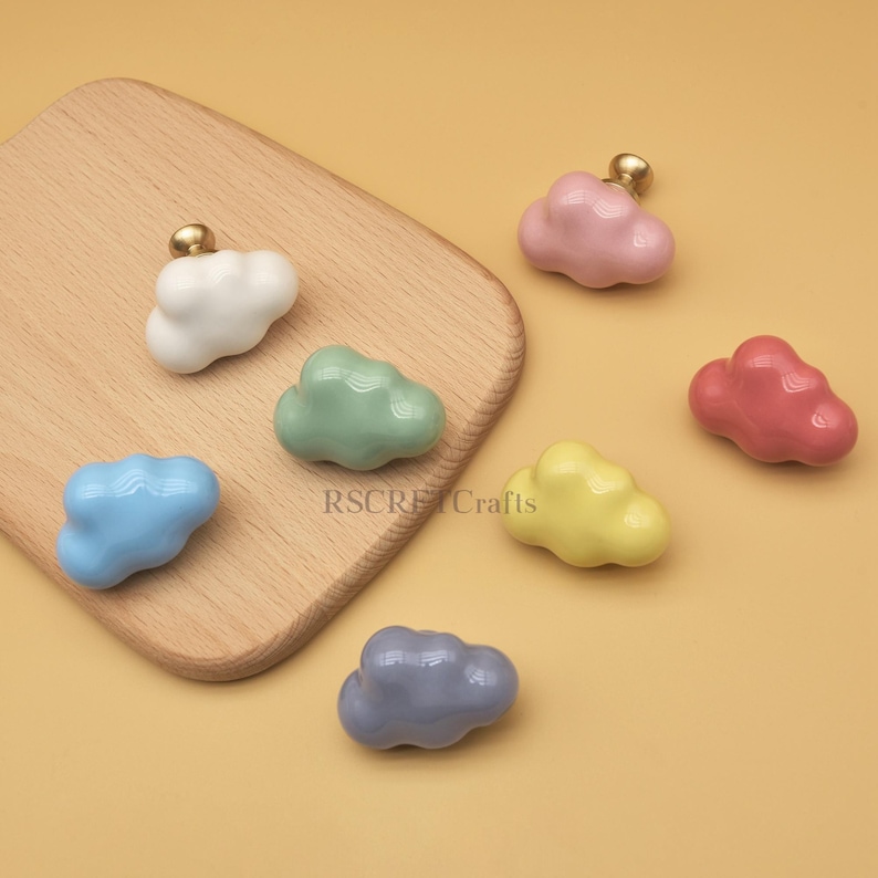 May include: A collection of cloud-shaped cabinet knobs in various colors, including white, pink, red, yellow, blue, green, and gray. Each knob has a gold-colored stem. The knobs are displayed on a wooden cutting board and a yellow background.