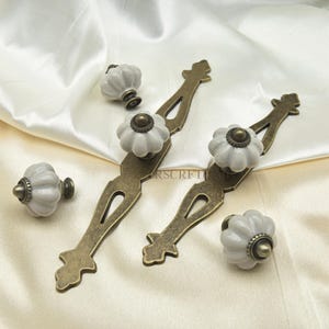 Może przedstawiać: Zestaw okuć do szafek, w tym dwa uchwyty ze starego brązu i cztery białe, ceramiczne gałki w kształcie dyni. Okucia są ułożone na kremowej satynie, eksponując detale dekoracyjne.