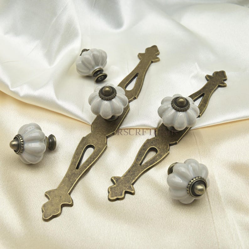 Może przedstawiać: Zestaw okuć do szafek, w tym dwa uchwyty ze starego brązu i cztery białe, ceramiczne gałki w kształcie dyni. Okucia są ułożone na kremowej satynie, eksponując detale dekoracyjne.