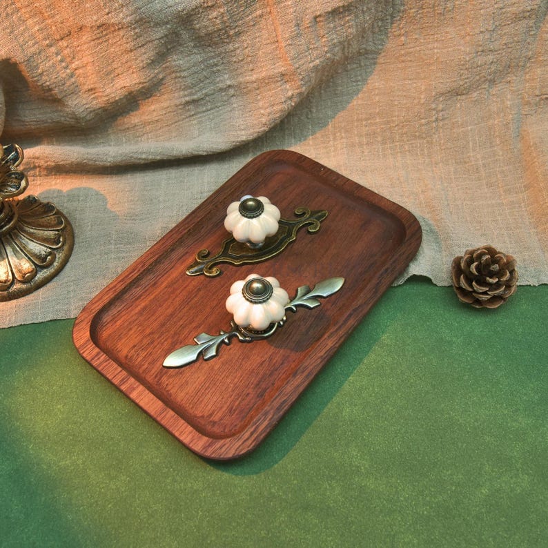 Może przedstawiać: Drewniana taca do serwowania z dwoma dekoracyjnymi uchwytami do szuflad. Uchwyty do szuflad mają białe ceramiczne gałki z brązowymi metalowymi akcentami. Szyszka sosnowa leży na zielonej powierzchni, a w tle znajduje się beżowa tkanina.