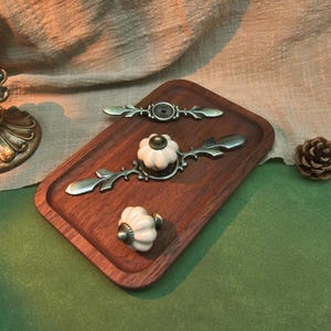 Może przedstawiać: Kolekcja okuć do szafek w stylu antycznym, w tym dwa ozdobne uchwyty i dekoracyjny gałek. Uchwyty mają brązowe wykończenie z detalami w kształcie liści, a gałka ma biały, kwiatowy wz&oacute;r.