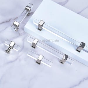 May include: A collection of clear acrylic cabinet pulls with polished silver-tone hardware. The pulls are rectangular and cylindrical, with a modern design. The image is on a white and marble background. The text "RSCRFTCrafts" is visible.