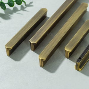 May include: Close-up of several antique brass cabinet pulls. The pulls are rectangular with rounded edges and a slightly curved handle. The finish is a warm, aged brass colour. The text "RSCRFT" is visible on one of the pulls.