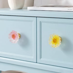 May include: A light blue cabinet featuring two drawers, each equipped with a decorative flower-shaped knob. The left knob is translucent pink with a textured center, while the right knob is translucent yellow with a textured center.