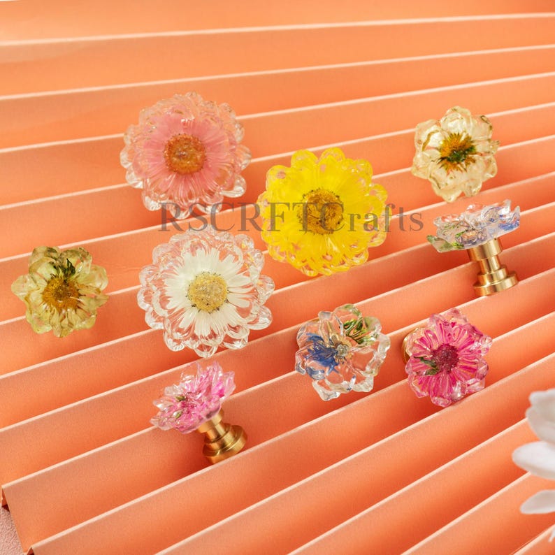 May include: A collection of round cabinet knobs featuring embedded dried flowers in clear resin with gold-toned metal bases. The floral designs include pink, yellow, white, and blue petals. The text RSCRF Crafts is visible in the center.
