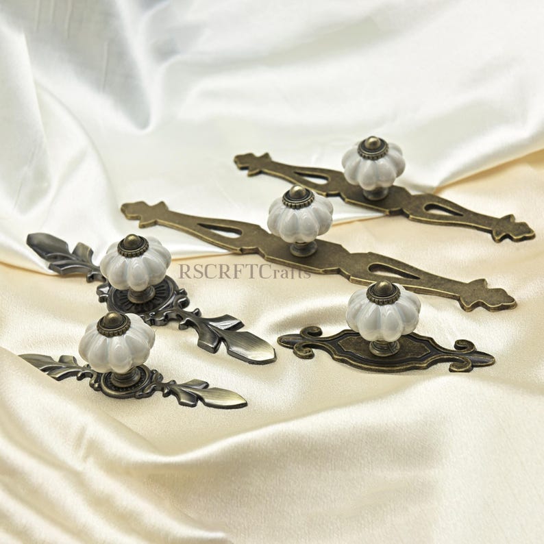 Może przedstawiać: Kilka uchwyt&oacute;w do szafek w stylu antycznym z białymi ceramicznymi gałkami i ozdobnymi, brązowymi płytkami tylnymi ułożonych na kremowej satynowej powierzchni. Uchwyty r&oacute;żnią się wzornictwem, niekt&oacute;re z akcentami w kształcie liści.