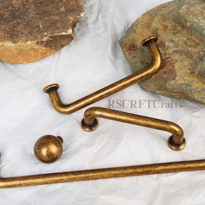 May include: A collection of antique bronze cabinet hardware, including handles and a knob. The handles have a curved design with rounded ends. The hardware is displayed on a white surface with natural stone accents. The text "RSCFTCrafts" is visible.