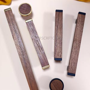 May include: A selection of wooden cabinet hardware, including knobs and pulls. The knobs have a circular design with a dark wood centre and a gold-coloured rim. The pulls are rectangular with dark wood and gold or black accents.