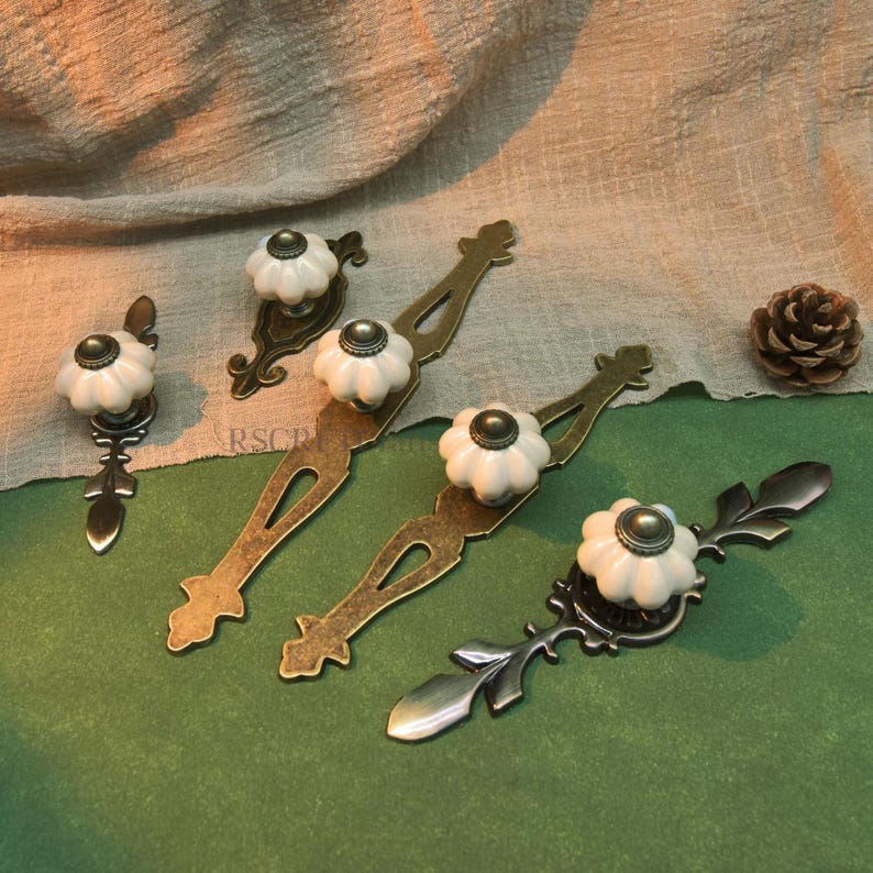 Może przedstawiać: Kolekcja uchwyt&oacute;w i gałek do szafek w stylu antycznym. Gałki są białe z ciemnym środkiem, a uchwyty mają ozdobne metaloplastyki w odcieniach brązu i ciemnego srebra. Obecna jest r&oacute;wnież szyszka.