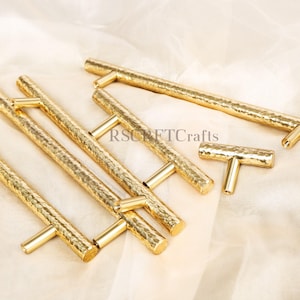 Puede incluir: Un conjunto de tiradores dorados para armarios con textura martillada. Los tiradores tienen forma de T con una barra rectangular y un asa cilíndrica. El hardware está dispuesto sobre una tela color crema.