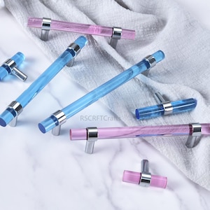 Może przedstawiać: Uchwyty do szafek w odcieniach niebieskiego i różowego, o przezroczystym, cylindrycznym kształcie i srebrnych metalowych akcentach. Uchwyty są ułożone na białej marmurowej powierzchni i szarej tkaninie. Wymiary w centymetrach.