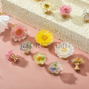 May include: A collection of decorative drawer knobs featuring clear resin tops embedded with various dried flowers in shades of pink, yellow, white, and blue, each attached to a gold-toned metal base. The text "SECRET Crafts" is visible.