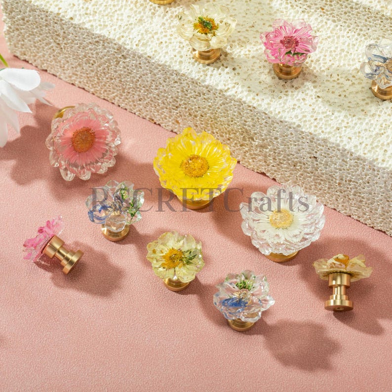 May include: A collection of decorative drawer knobs featuring clear resin tops embedded with various dried flowers in shades of pink, yellow, white, and blue, each attached to a gold-toned metal base. The text "SECRET Crafts" is visible.