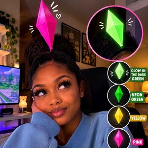 May include: A headband featuring a diamond-shaped plumbob topper. The topper is available in glow-in-the-dark green, neon green, yellow, pink, and red. The image displays the pink topper on a headband and includes text stating FREE GIFT INCLUDED.