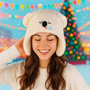 May include: A cream-colored winter hat with bear ears and an embroidered koala face. The hat has a black nose, closed eyes, and a blue tongue. The background features a Christmas tree and colorful bunting.