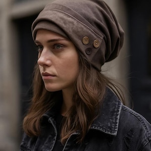 Può includere: Un berretto marrone con tre bottoni decorativi sul lato. Il cappello è realizzato in un materiale morbido e ha un bordo ripiegato. Il cappello è di colore neutro e può essere indossato con molti outfit.