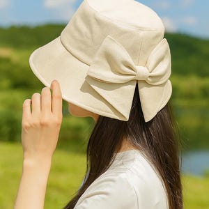Könnte beinhalten: Ein cremefarbener Sonnenhut mit breiter Krempe und einer großen Schleife an der Seite. Der Hut ist aus einem strukturierten Stoff gefertigt und wird von einer Person im Freien getragen. Der Hintergrund zeigt grünes Gras und einen blauen Himmel.