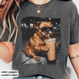 Może przedstawiać: Ciemnoszary t-shirt z nadrukiem graficznym przedstawiającym brązowego psa w czarnych okularach przeciwsłonecznych, trzymającego przezroczysty plastikowy kubek z mrożoną kawą i słomką. Pies siedzi na szarej powierzchni.